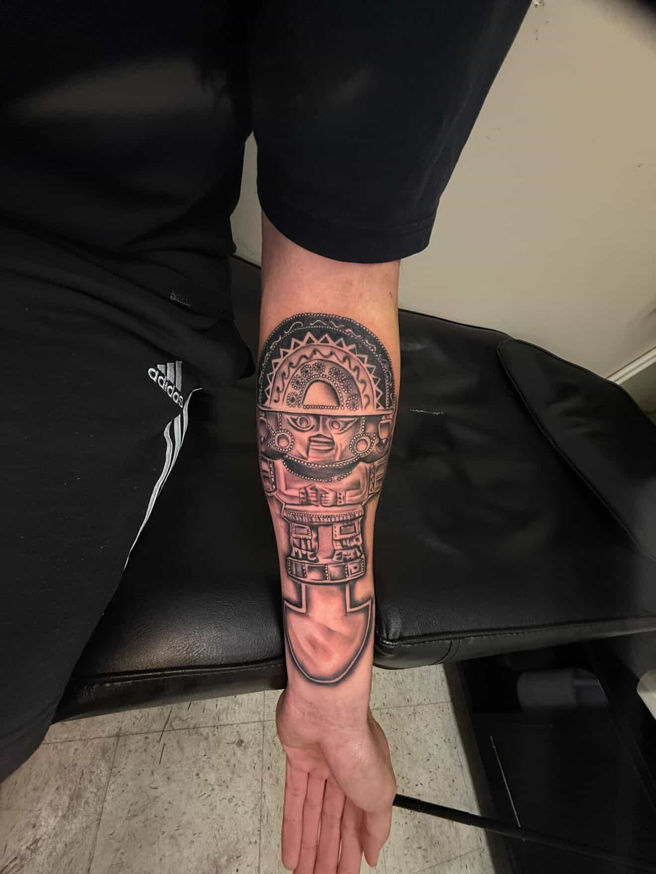 Black and gray Aztec warrior tattoo with detailed headdress and geometric patterns on forearm